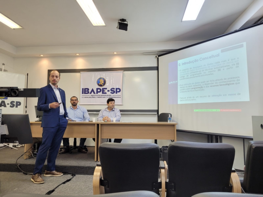 A imagem mostra Bruno Henrique Gazzinelli, palestrante e engenheiro civil, apresentando sobre raspagem de dados em um evento do IBAPE-SP. Ele está de pé, usando terno azul, falando para uma audiência com um slide projetado ao fundo. No slide, há informações sobre a introdução conceitual da raspagem de dados. Ao fundo, dois outros participantes estão sentados à mesa, e o banner do IBAPE-SP está pendurado na parede.